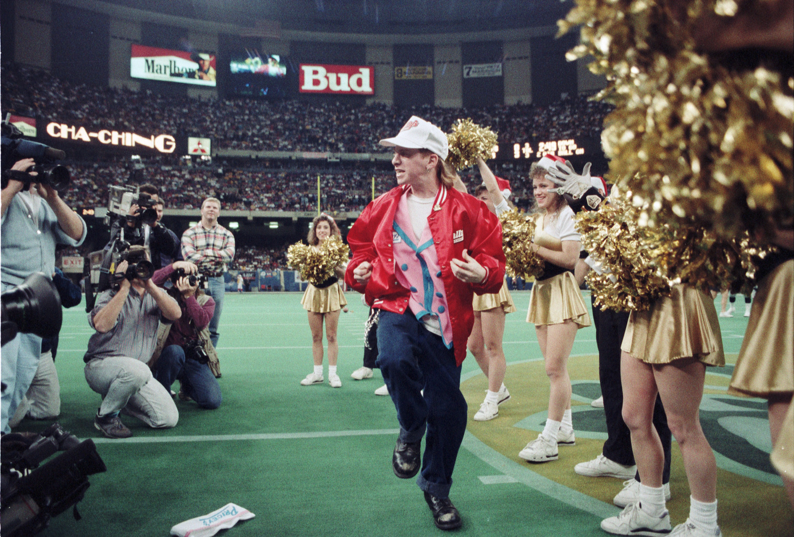 A man in a red jacket and baseball cap celebrates on a football field, surrounded by photographers and cheerleaders with gold pom-poms. A crowd fills the stadium in the background.
