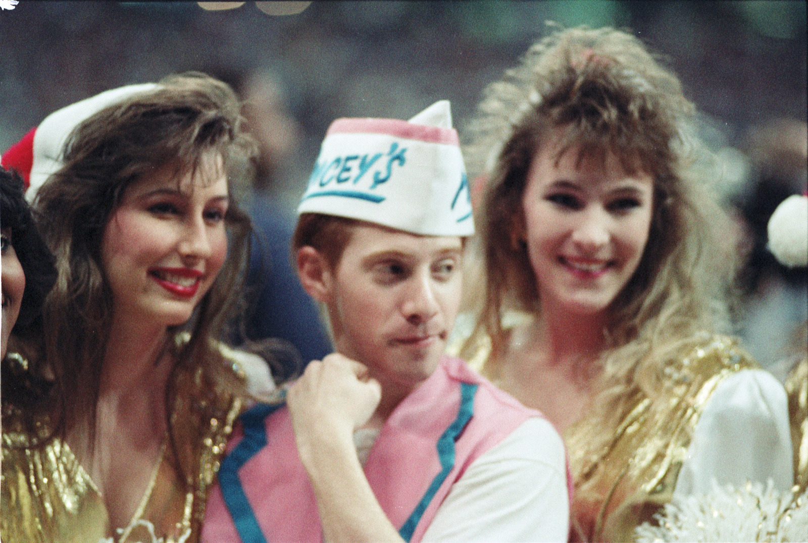 A person wearing a paper hat and pink and white outfit poses with two women in gold sequined dresses. Theyre all smiling in a lively, festive atmosphere.