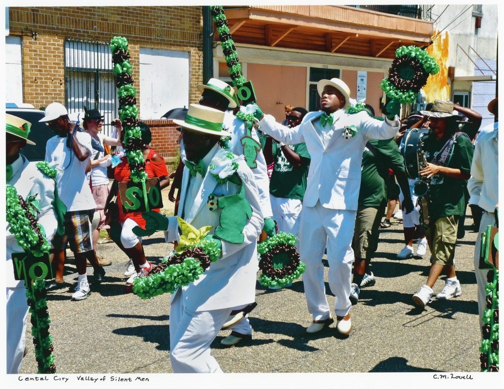 A vibrant parade scene with people dressed in white suits and green accessories. They hold decorative poles and wreaths. Onlookers stand along the street, enjoying the festive atmosphere in the bright sunlight.