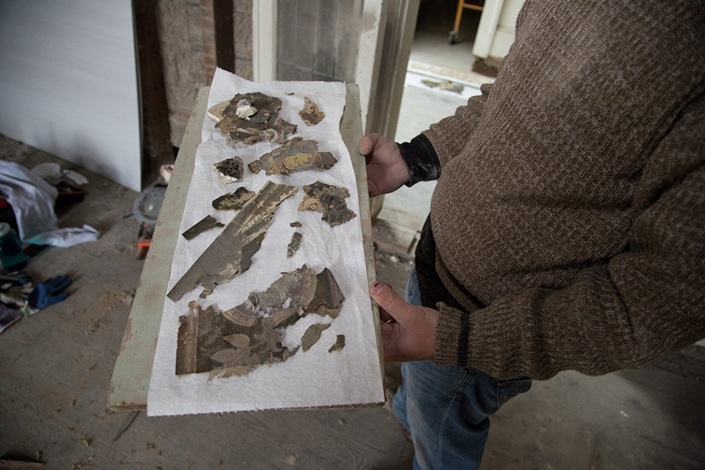 A person holds a rectangular tray covered with paper towels featuring several weathered and fragmented artifacts, possibly wallpaper remnants, while standing in a dimly lit, unfinished room.