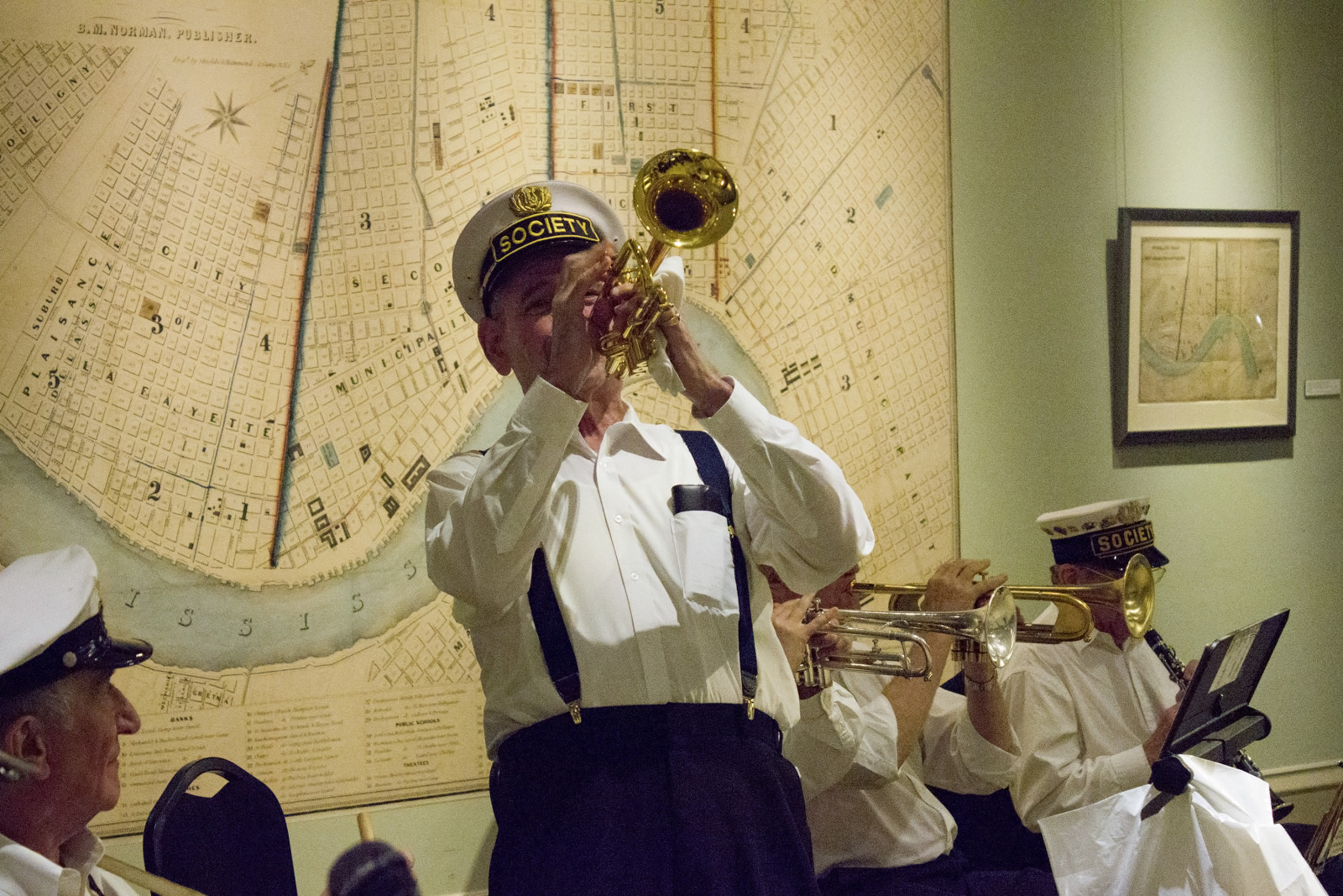 A brass band performing in front of an old, detailed map. The musicians are wearing white shirts, dark trousers, and hats. The lead trumpet player stands in the foreground, energetically playing his instrument.