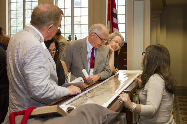 A group of people gather around a long table, examining a large map. One woman points at the map, while others look on with interest. Tall windows and a flag are visible in the background.