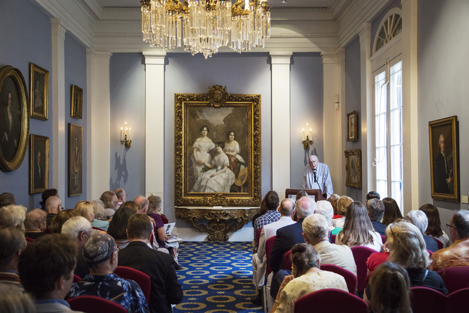 A crowded room with people seated, facing a speaker at a podium. The room is decorated with portraits on the walls, and a large, ornate painting of three women hangs prominently. A chandelier hangs from the ceiling, adding an elegant touch.