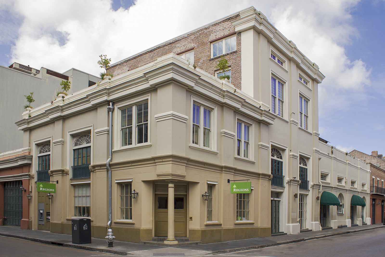 A corner view of a historic building with beige walls, arched windows, and green signs displaying Balconies. The ground floor has large windows and a door, while the upper floors feature French doors with small balconies. Cloudy sky above.