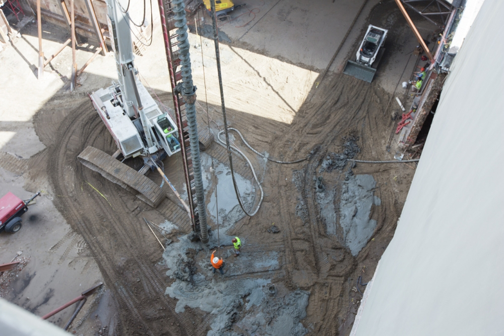 Aerial view of a construction site with workers in helmets and high-visibility clothing operating heavy machinery. Concrete is being poured into a deep foundation hole surrounded by steel supports and sandy ground.