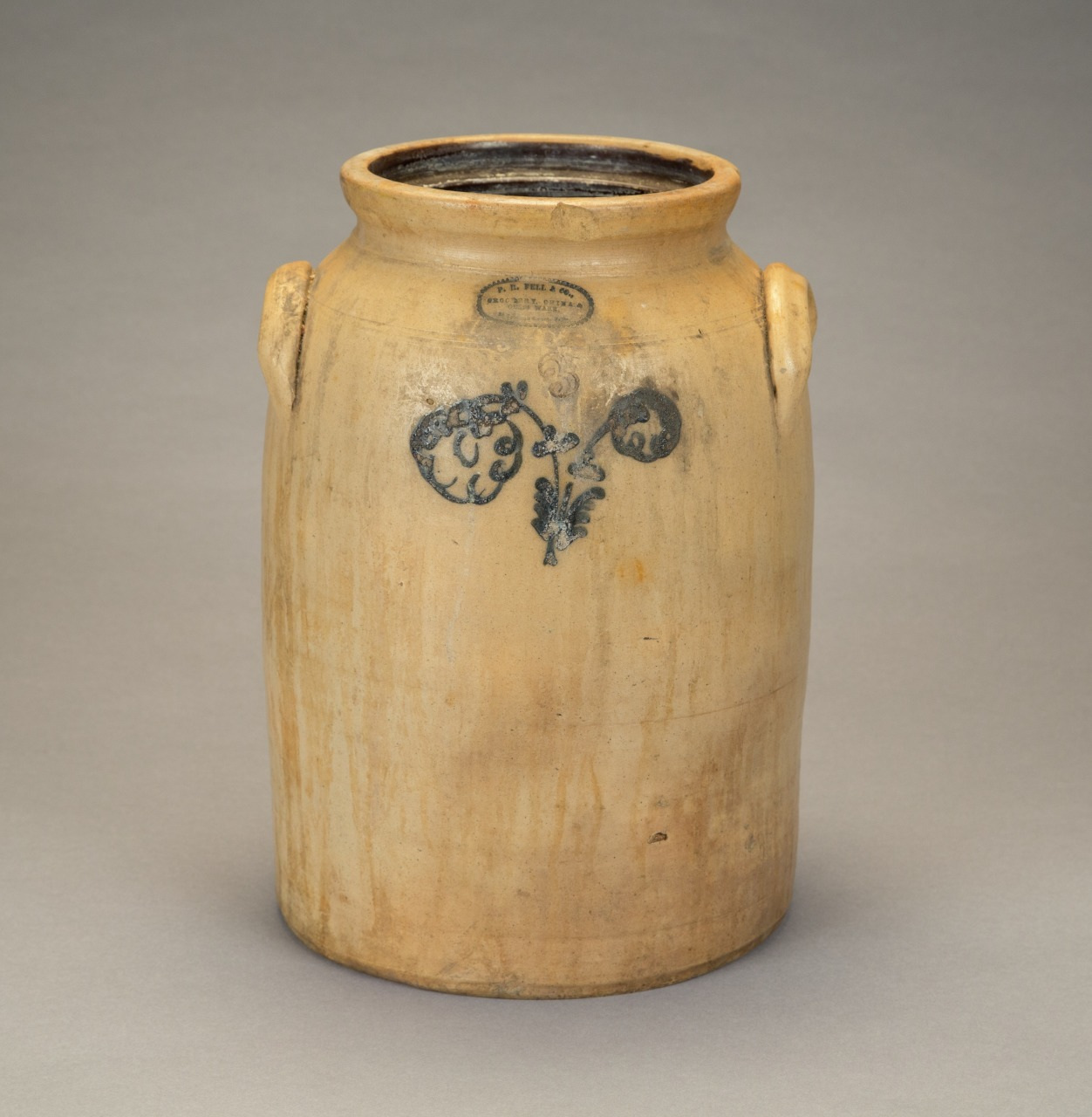 A vintage, beige stoneware crock with two small side handles and a blue floral design on the front, set against a neutral background.