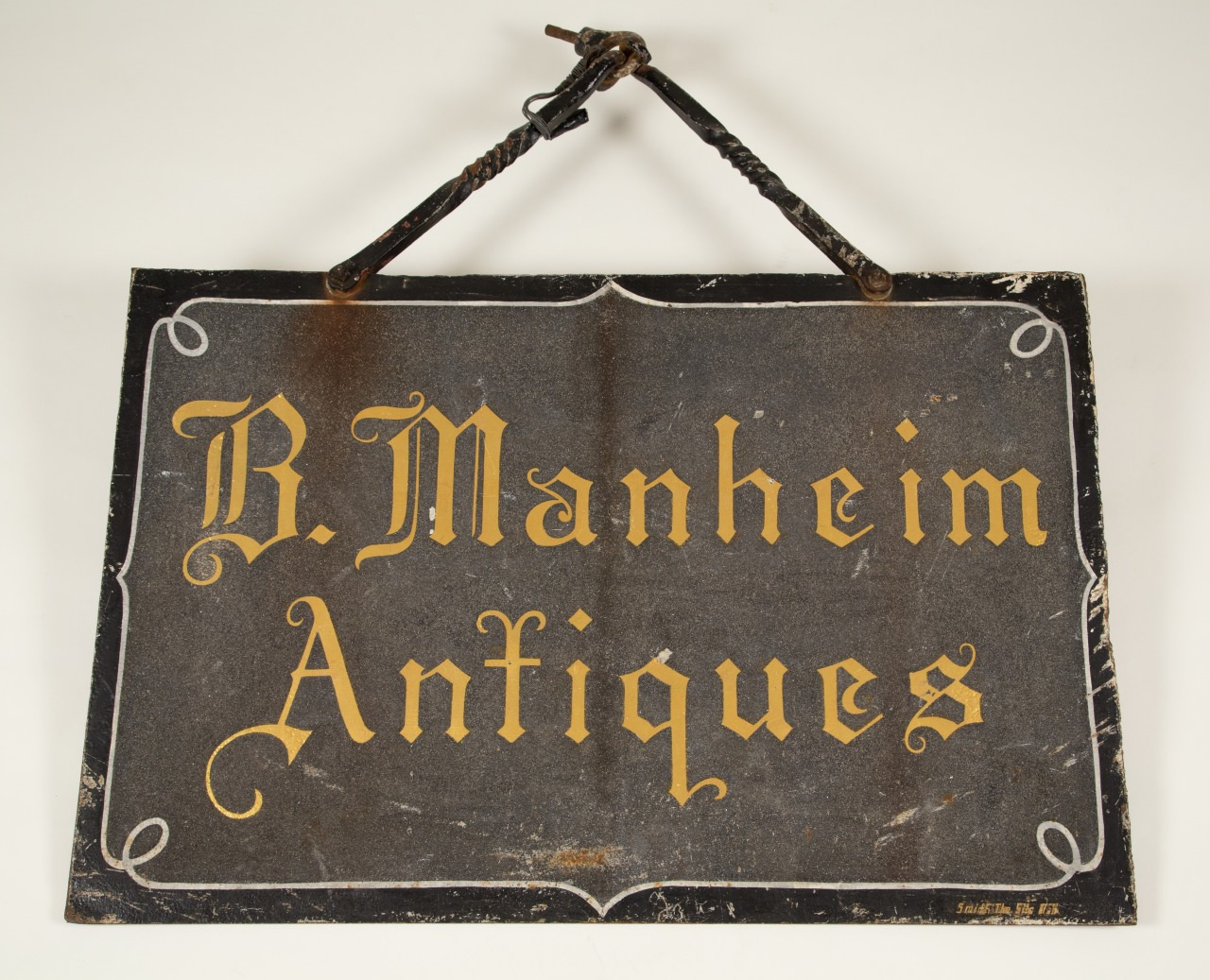 A vintage black sign with ornate yellow text reads “B. Manheim Antiques.” The sign has a weathered appearance with decorative corners and is hanging by a twisted metal handle.