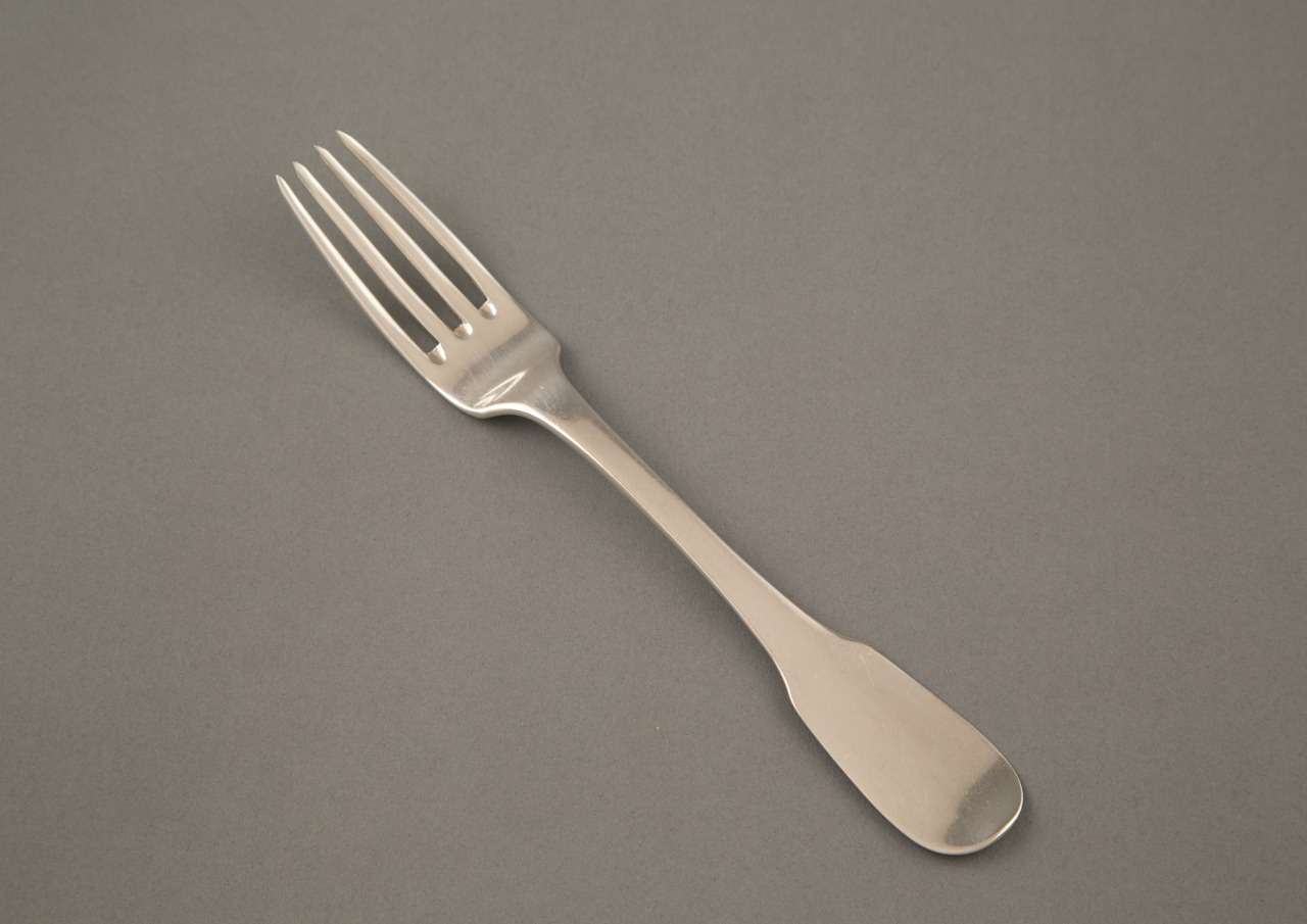 A stainless steel fork with a simple design lies on a flat, gray surface. The background is plain, highlighting the forks shiny, metallic appearance and traditional shape.