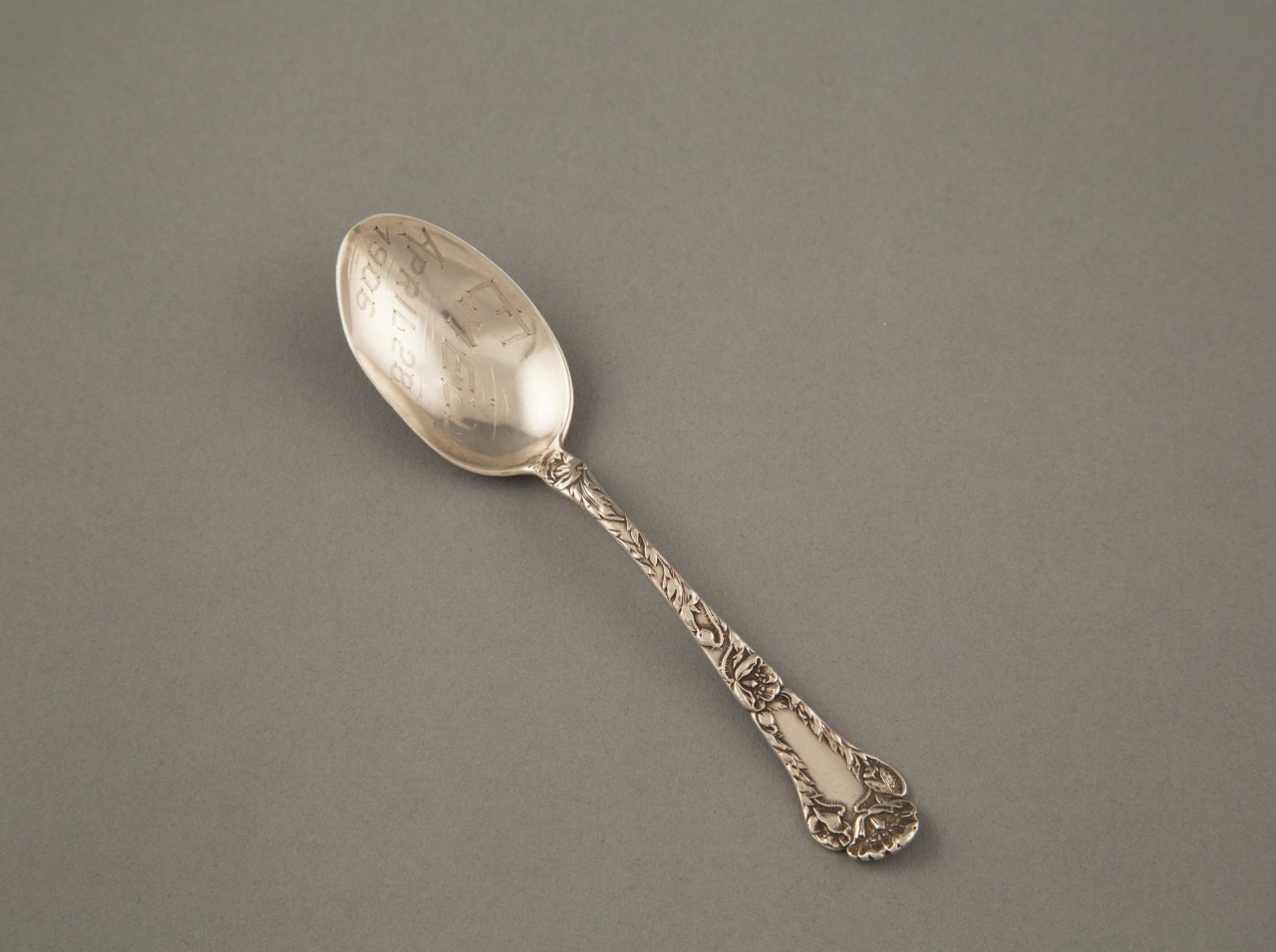 An ornate silver spoon with intricate floral designs on the handle, set against a plain grey background. The bowl of the spoon appears to have faint engravings.