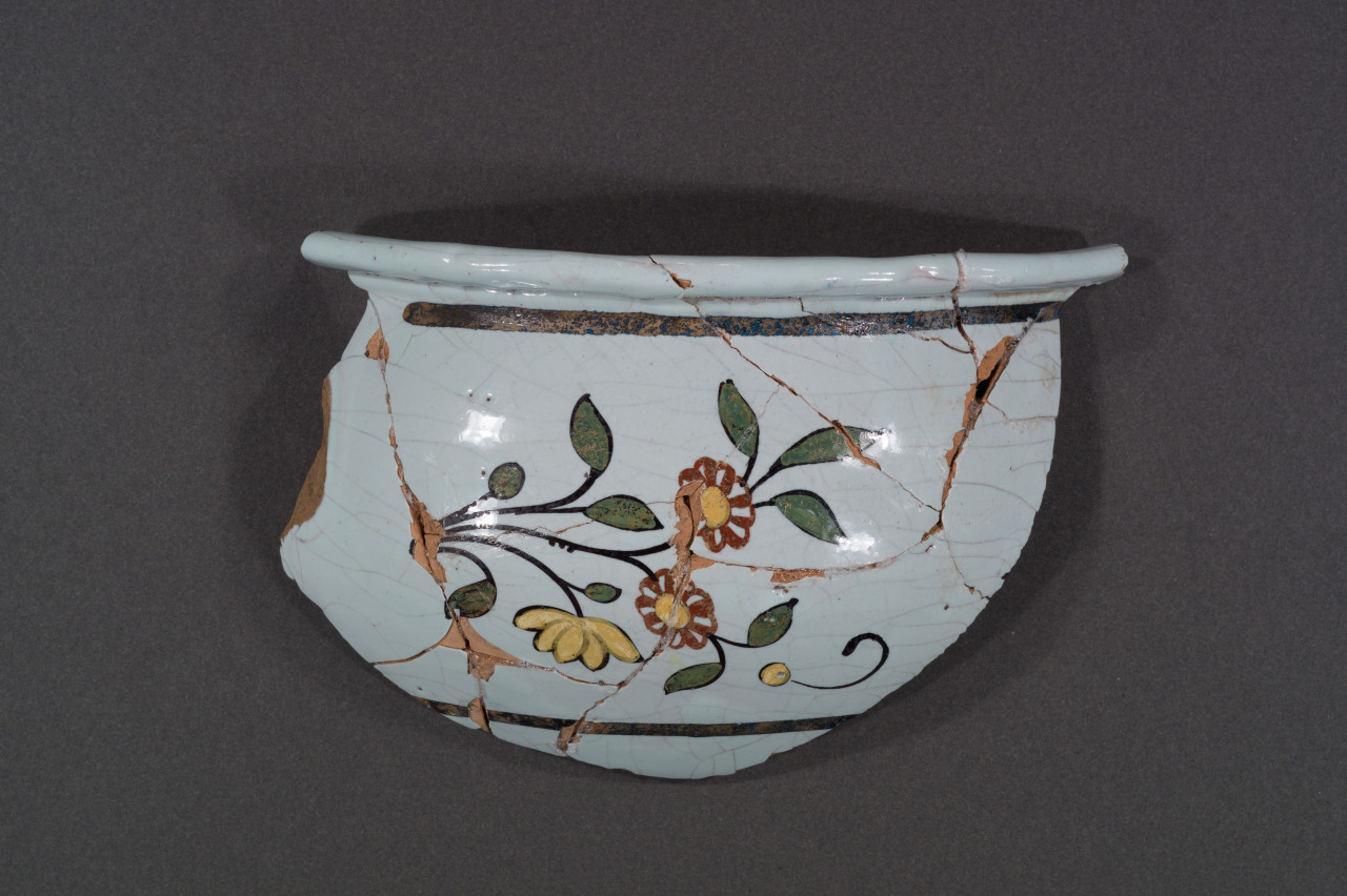A fragmented ceramic bowl with a light blue glaze and floral design in green, yellow, and brown. The bowl has visible cracks and missing pieces, against a plain gray background.