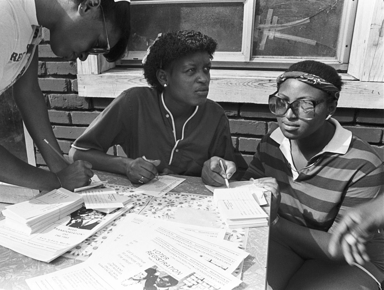 Three people are at a table with stacks of flyers and documents. Two are seated, looking at the camera, while one stands, writing. The scene appears to be a registration or sign-up event in an outdoor setting.