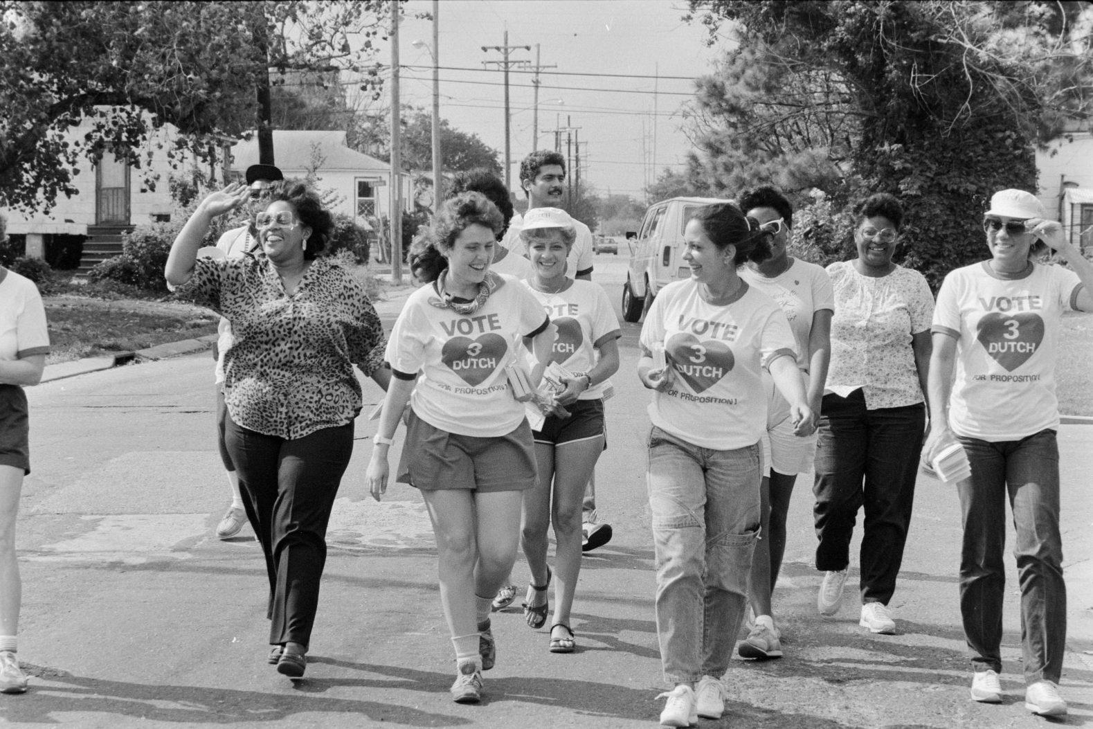 A group of people walking on a street, several wearing Vote 3 Dutch Morial shirts. Some are smiling and waving. Trees and houses are in the background. The mood appears lively and supportive.