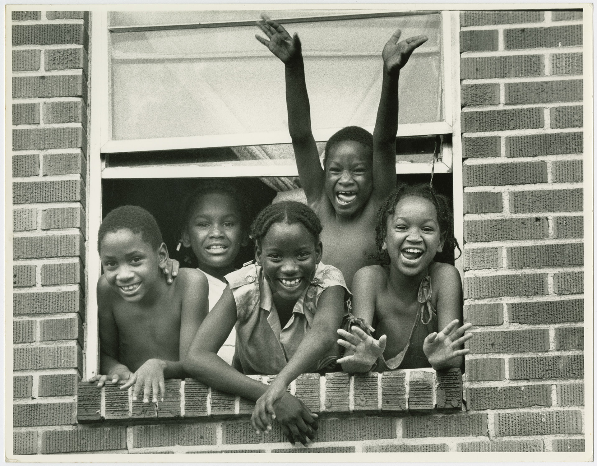 A black and white photo of five children smiling and waving out of the open window of a multistory brick building.