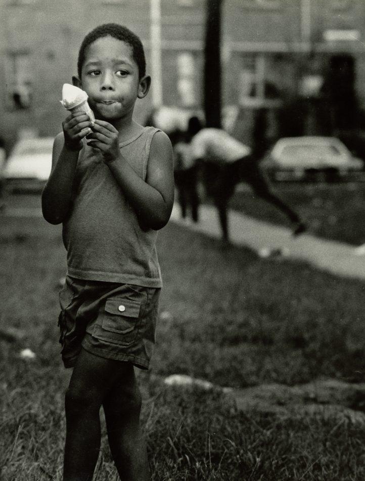 A young boy stands on grass, holding an ice cream cone while looking thoughtfully into the distance. Hes wearing a sleeveless top and shorts. In the background, there are blurred figures and parked cars on a street. The image is in black and white.