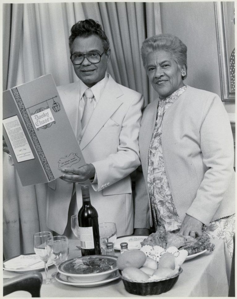 A man and woman stand together at a dining table. The man, wearing a light-colored suit, is holding a large packaged item. The table is set with wine, glasses, bread, and other dishes, with a curtain in the background.