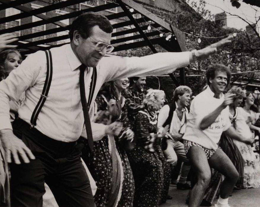 A group of people enthusiastically dancing outdoors under a pergola. The foreground features a man with glasses and suspenders striking a dynamic pose, while others around him join in the fun. Trees and a building are visible in the background.