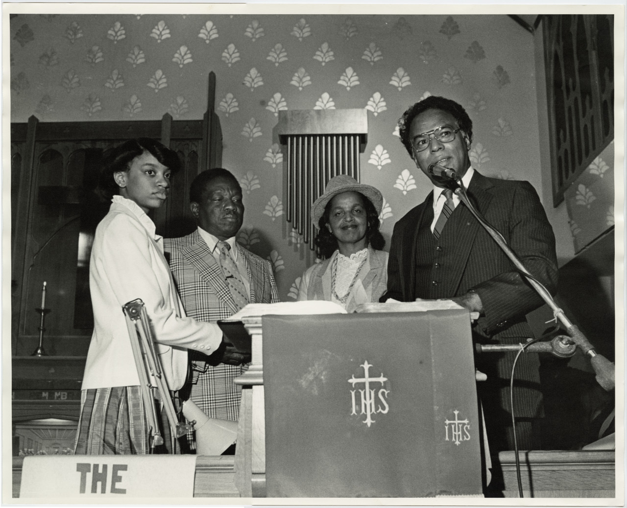 A man speaks at a podium with a microphone in a church, accompanied by two women and another man. They stand in front of pews and a patterned wall. The podium is draped with cloth featuring religious symbols.