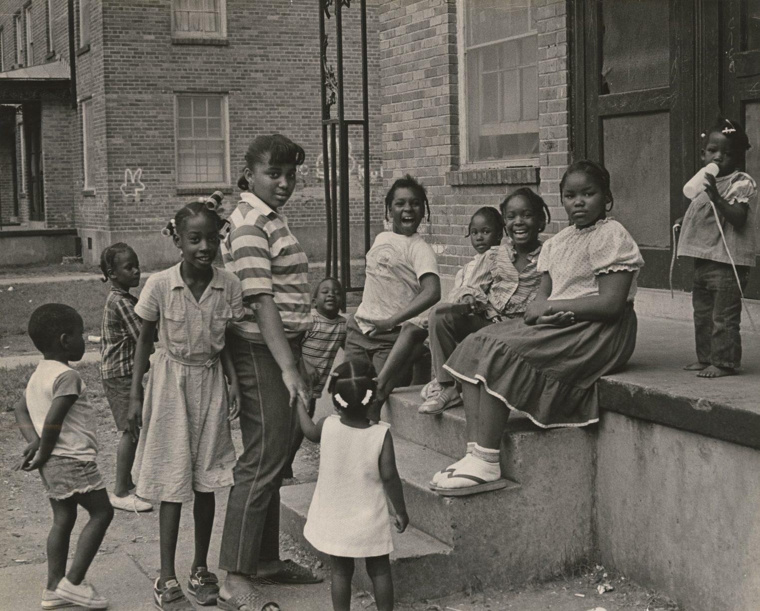 A group of children and a woman gather on the steps outside a brick building. The children are playing and smiling, with some sitting while others stand. One girl is holding a jump rope. The scene conveys a sense of community and camaraderie.