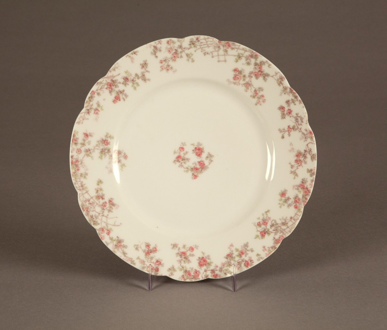 A delicate white porcelain plate with a scalloped edge, adorned with a pattern of small pink roses and green leaves around the rim and a cluster of roses in the center. The plate is displayed on a stand.