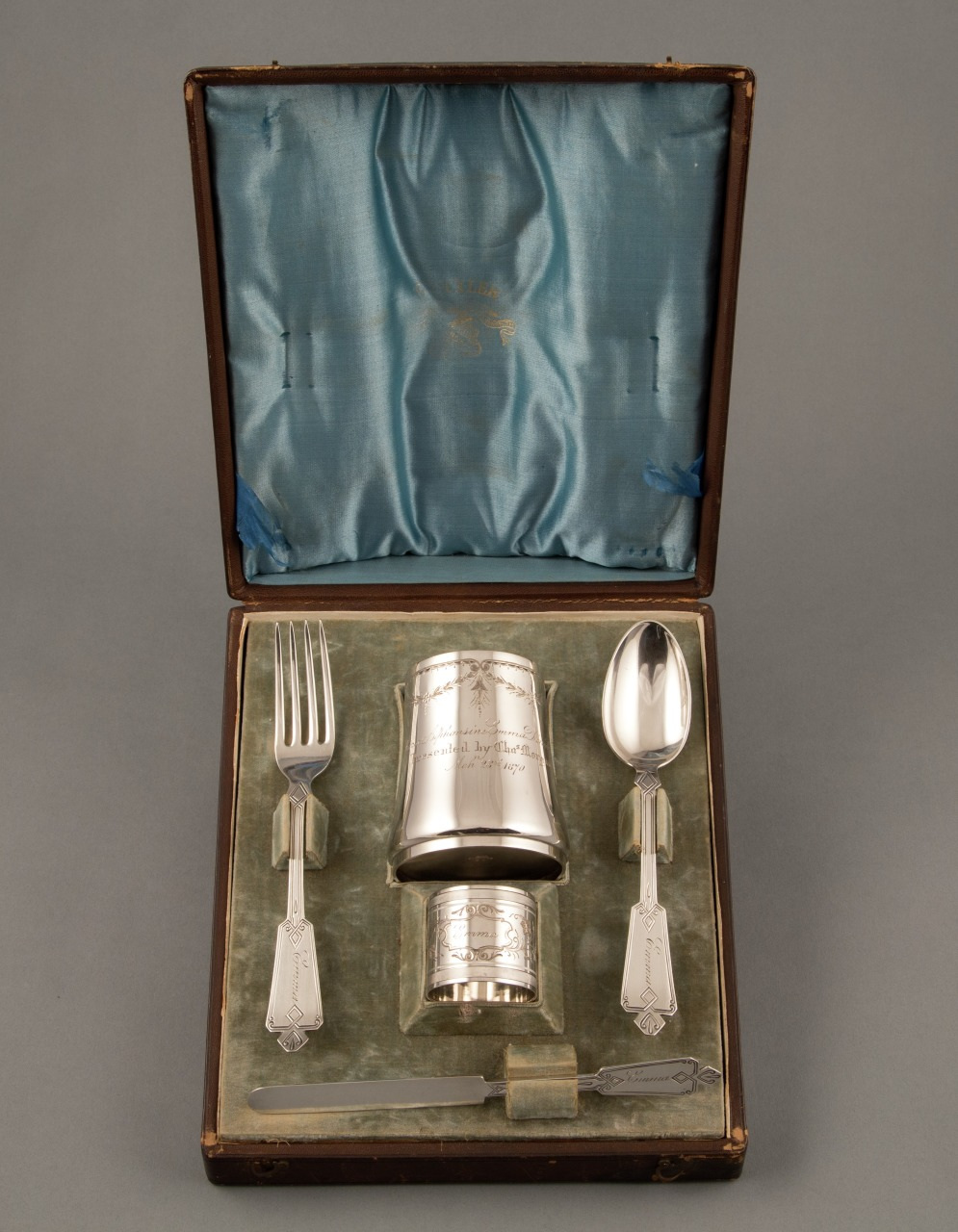 A vintage silver dining set in a wooden box with satin lining. Includes a fork, spoon, cup, napkin ring, and knife with ornate handles, arranged neatly on a padded base. The inside lid is lined with blue satin fabric.
