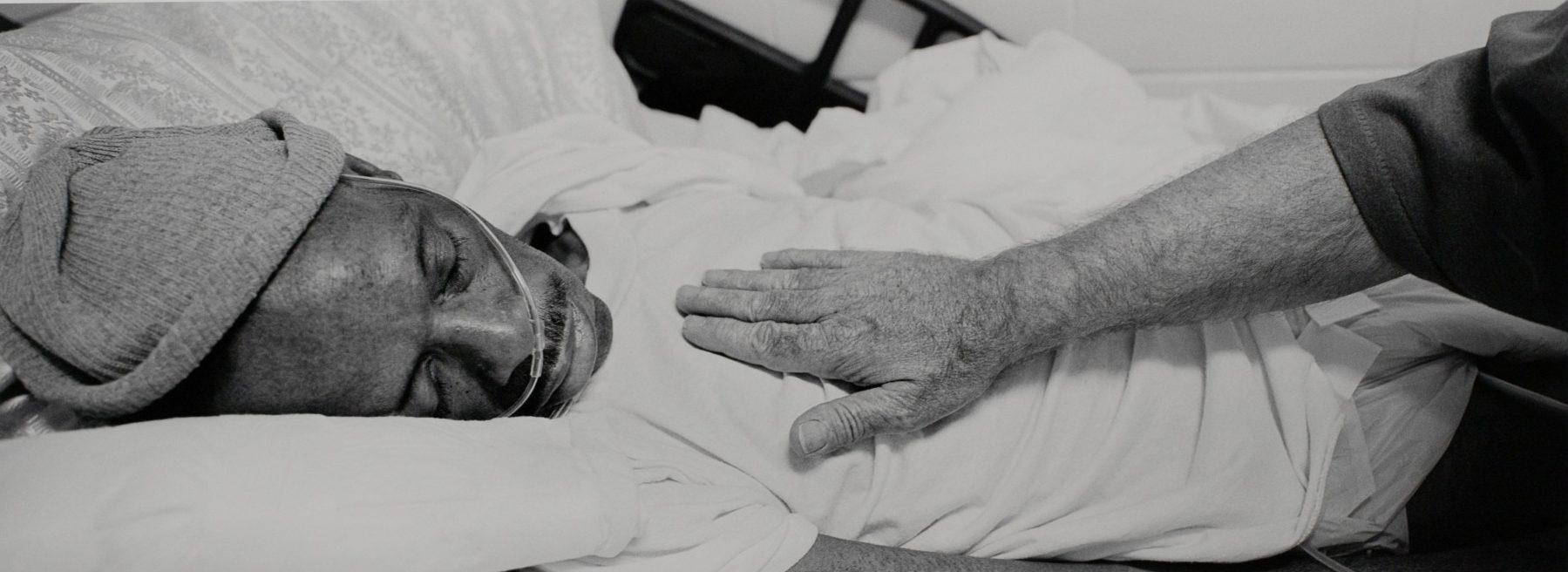 A black and white image of a person lying in a hospital bed wearing a nasal oxygen tube. They are covered with a blanket. A hand is gently placed on their chest, indicating care or examination.