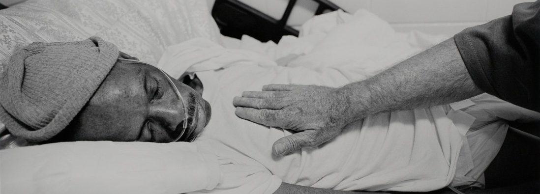 A person lies on a bed, wearing a beanie and a white shirt, with their eyes closed. Another persons hand rests gently on the lying persons chest, creating a comforting scene. The image is in black and white.