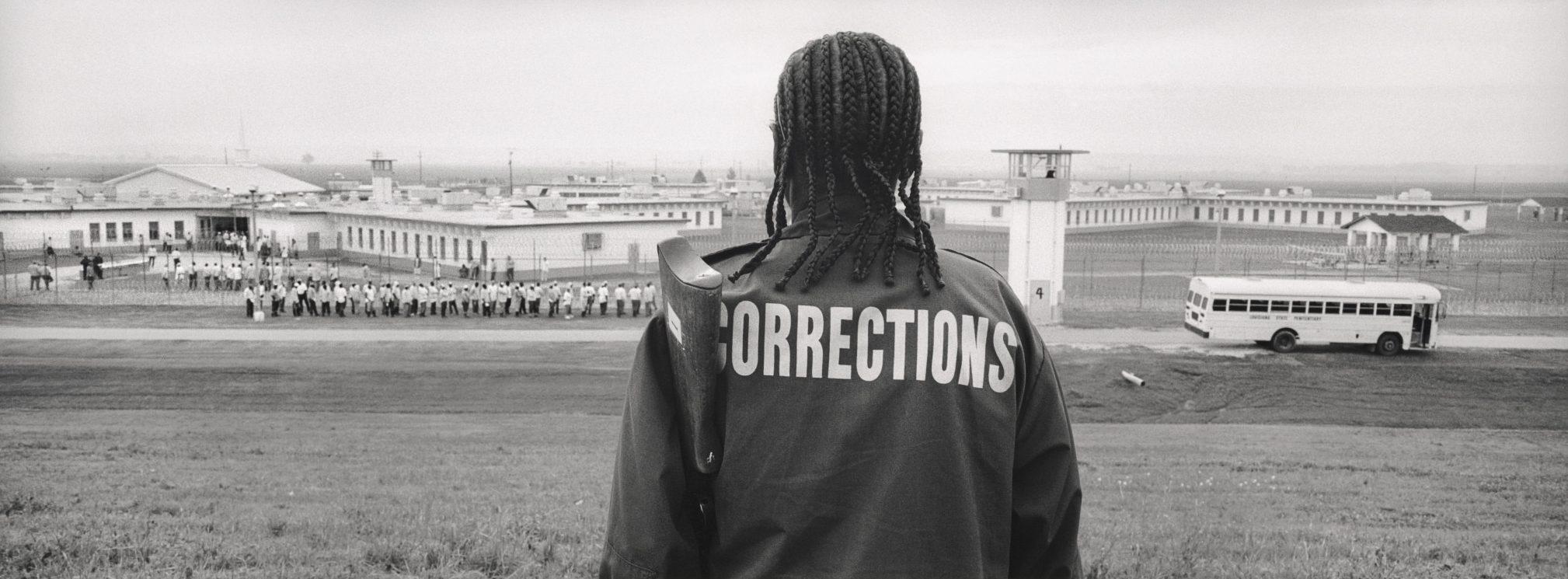 A person with braided hair, wearing a jacket labeled CORRECTIONS, stands on a grassy hill overlooking a prison complex. A group of people and a bus are visible in the background.