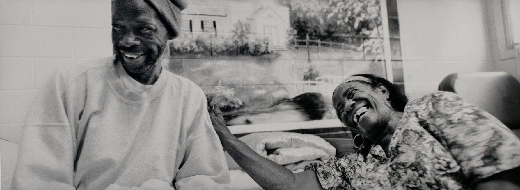 A black and white photo of a man and a woman laughing together. The man is wearing a beanie and sweatshirt, and the woman is touching his shoulder, wearing a floral blouse. They are indoors with a painted mural of a house in the background.