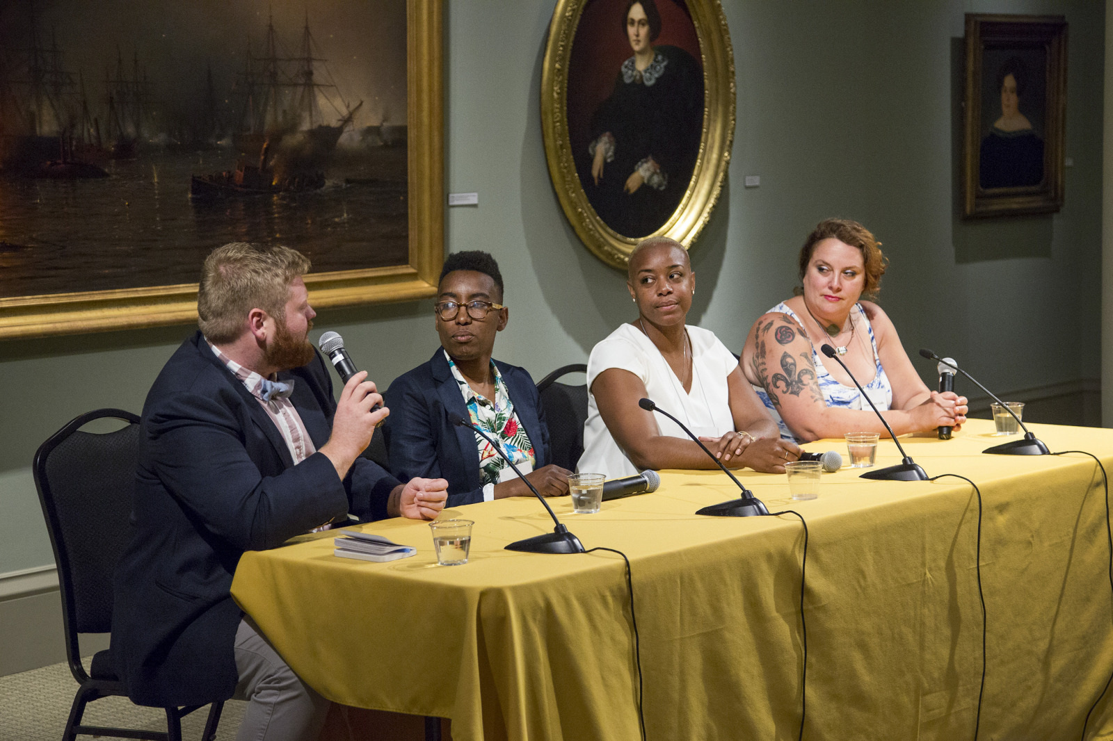 A panel of four people sitting at a table with microphones discuss in a room adorned with paintings. The man on the left, holding a microphone, is speaking while the others listen attentively.