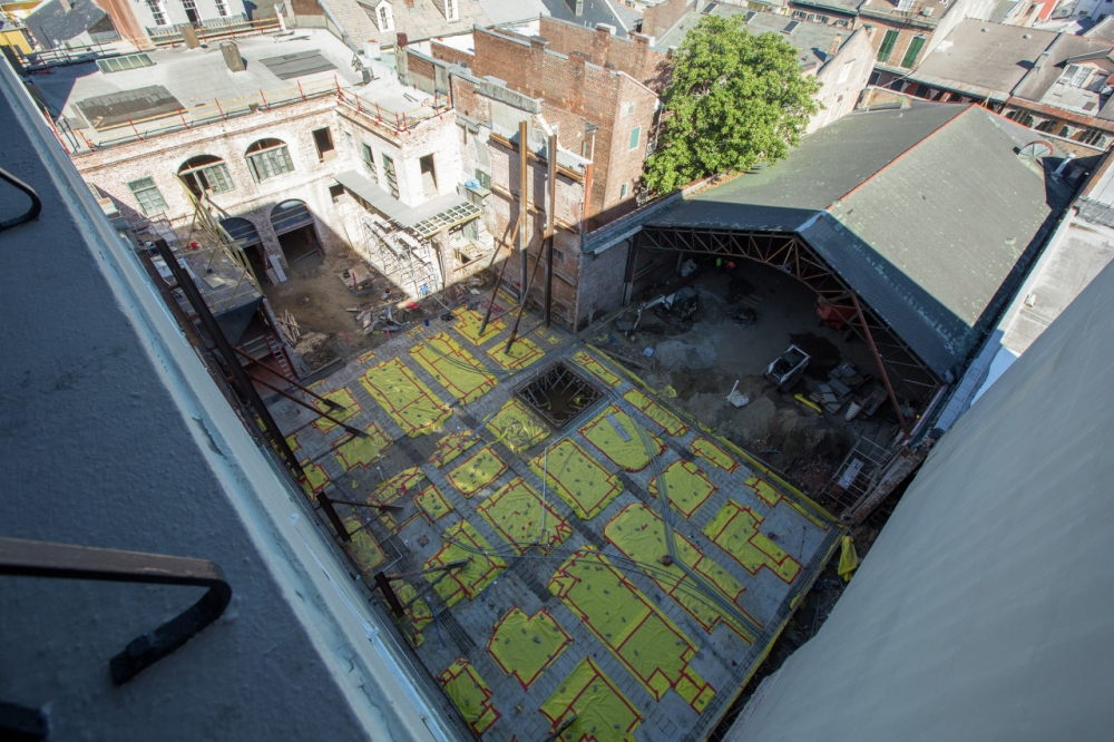 Aerial view of an urban construction site with steel beams and a grid pattern on the ground. Surrounding buildings and a partial metal roof frame are visible. A tree stands near the site in an otherwise industrial setting.