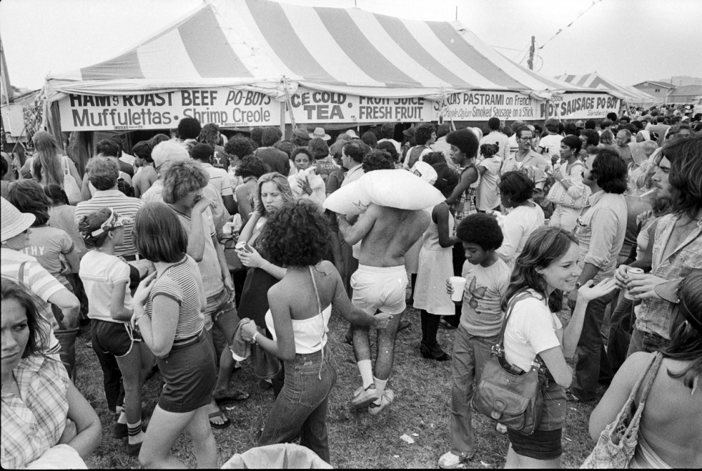 A busy outdoor food festival with a striped tent in the background. Diverse people gather, some holding drinks. A man with a pillow on his shoulder stands out. Signs advertise roast beef, poboys, muffulettas, and other foods.