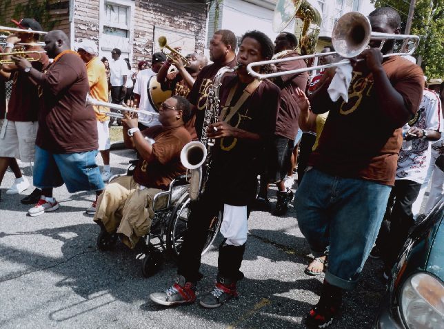 A lively street band performs during a parade. Musicians play brass instruments, with a man in a wheelchair playing saxophone. They wear matching brown shirts and are surrounded by spectators. The scene is vibrant and energetic.