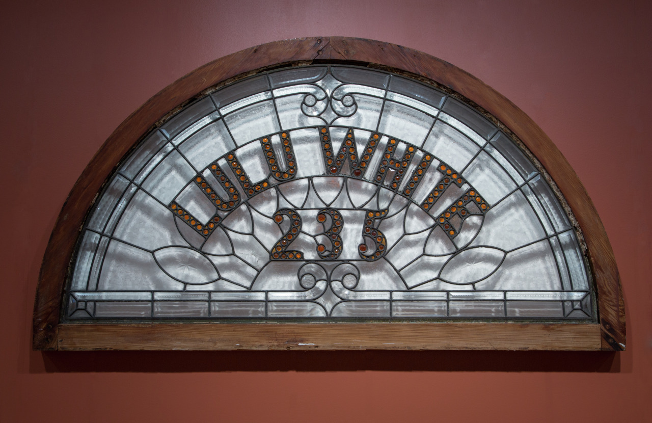 A semicircular stained glass window with the words LULU WHITE 235 in decorative lettering. The glass design features geometric patterns, framed by a wooden arch against a reddish-brown background.