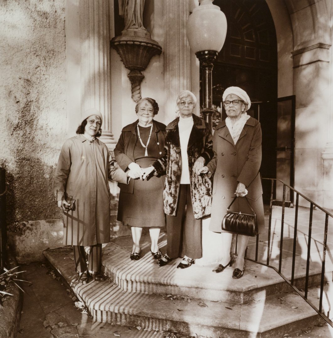 Four women stand on the steps of a cathedral, wearing Sunday clothes and hats.