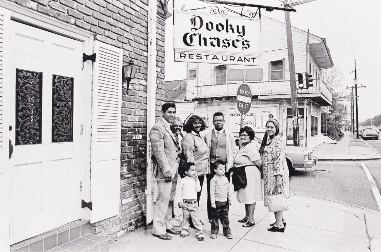 A black and white photo of a group of people standing in front of a brick building with a sign reading Dooky Chases Restaurant. The group includes adults and children, all dressed in formal attire. A street with cars and buildings is in the background.