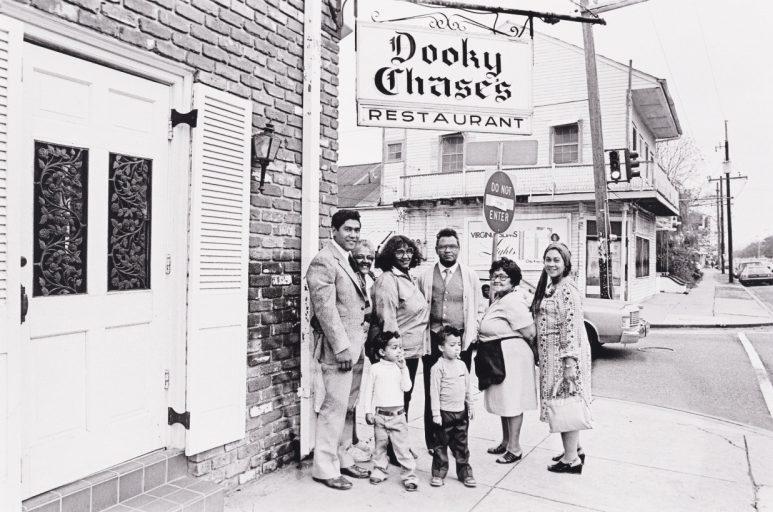 A group of adults and children stand outside Dooky Chases Restaurant. They smile at the camera in front of the restaurants sign, with a brick wall and street in the background. The scene captures a moment of gathering and joy.