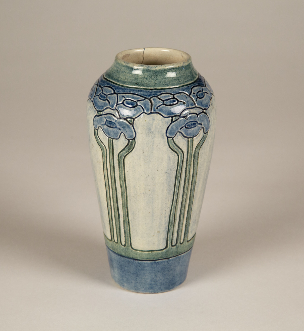 A ceramic vase with a blue and cream glaze featuring stylized floral patterns in blue near the top. The vase has vertical green lines extending from the base of the flowers. The overall shape is cylindrical with a slightly narrower neck.
