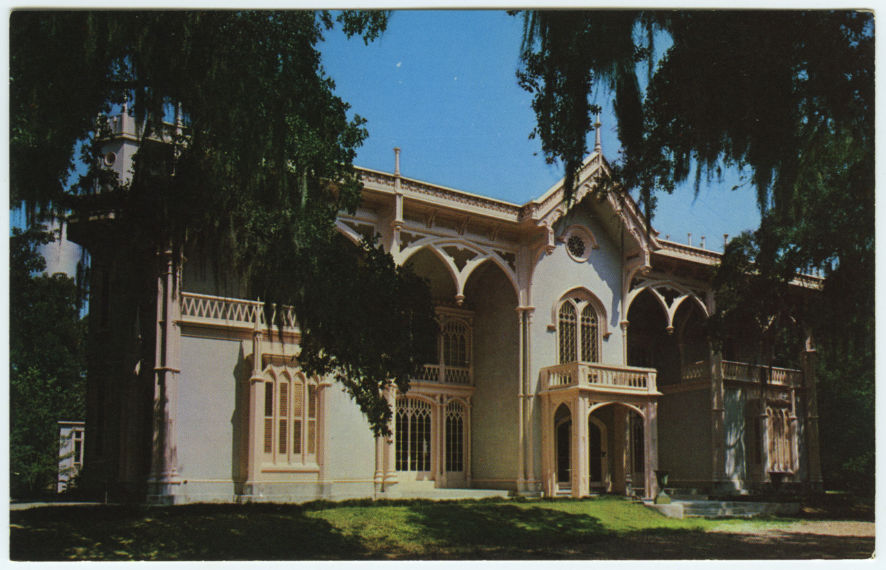 A grand historic mansion with intricate Gothic-style architecture, featuring arched windows and detailed woodwork, set against a clear blue sky. Large trees with hanging moss frame the scene, providing shade and enhancing the serene atmosphere.