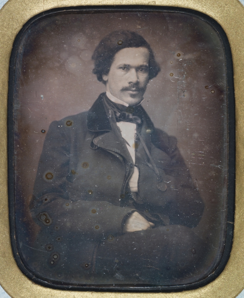 A vintage photograph of a man with wavy hair and a mustache, dressed in a 19th-century style suit with a bow tie. He is sitting and looking forward, with a neutral expression. The image has a sepia tone and shows signs of aging.