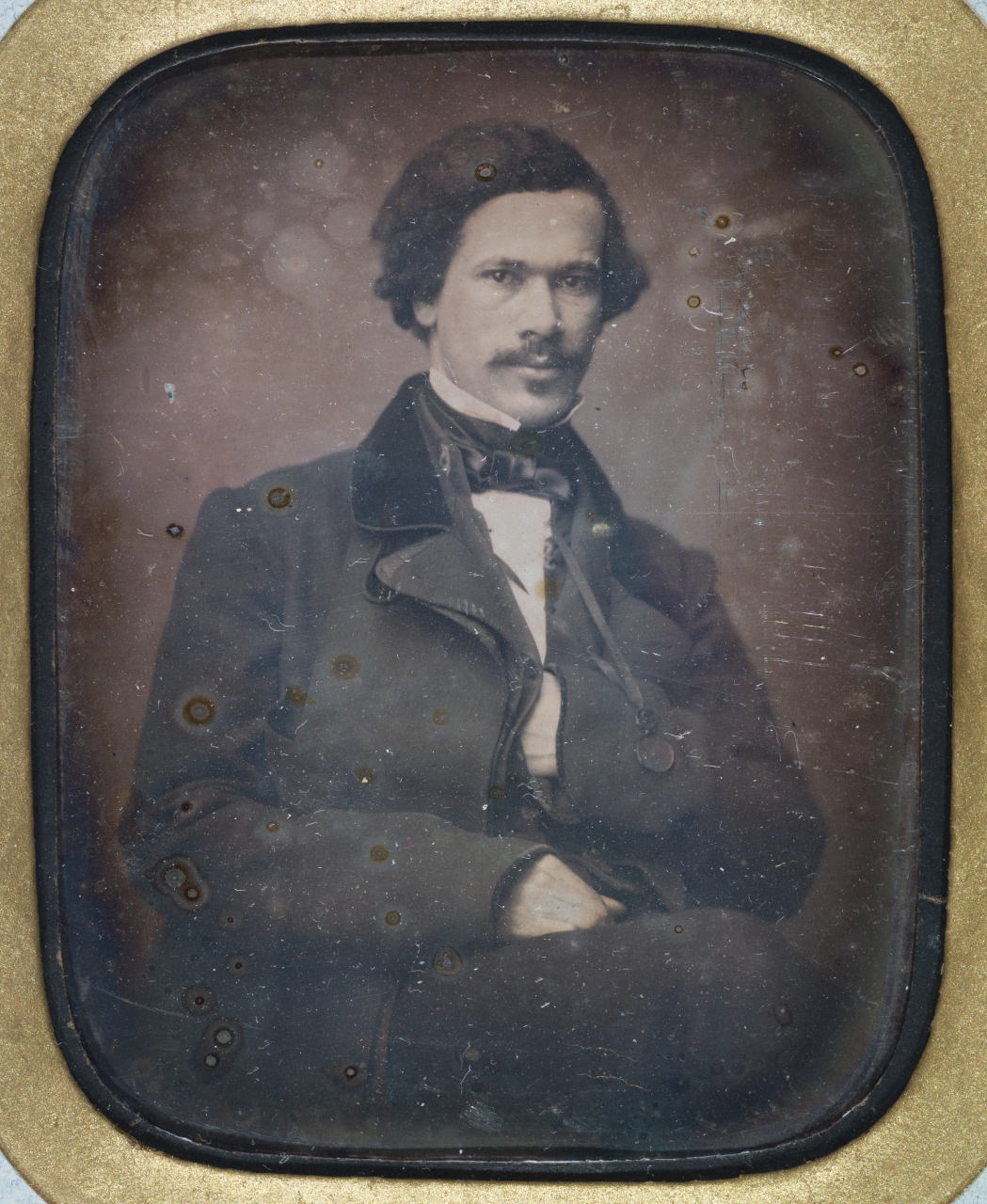 A vintage black-and-white portrait of a man in 19th-century attire. He is seated, wearing a dark coat and a bow tie, with a neutral expression. The photograph has an aged, antique appearance with a gold-toned frame.