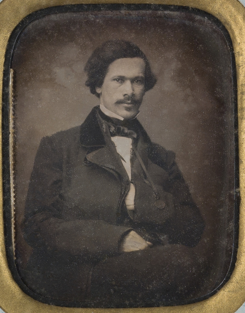 A vintage photograph of a man with a mustache, wearing a dark suit and bowtie. He is seated, facing forward, and has a calm expression. The image has an ornate golden frame, characteristic of historical portrait photography.