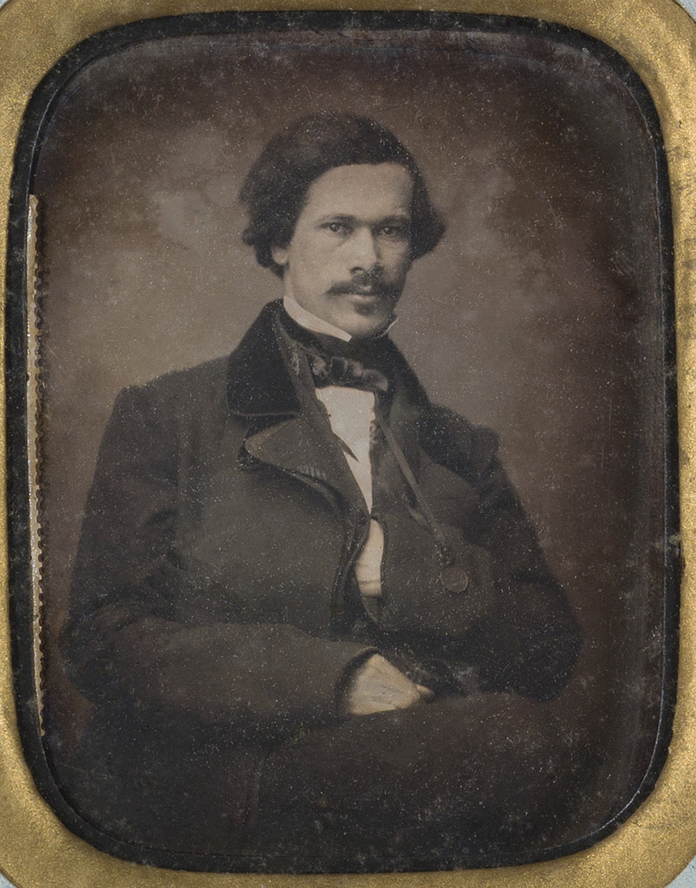 A vintage portrait of a man with a mustache and dark hair, dressed in a suit with a bow tie. He sits with his arms crossed, looking directly at the camera. The image is framed in an antique gold-bordered case.