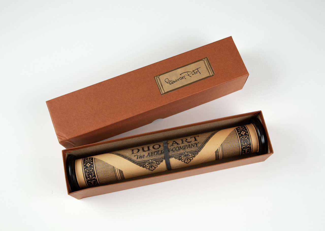 A brown box open to reveal a scroll labeled Duo-Art, The Aeolian Company. The scroll features ornamental designs and is nestled in the box with a label on the lid. The overall presentation is elegant and vintage.