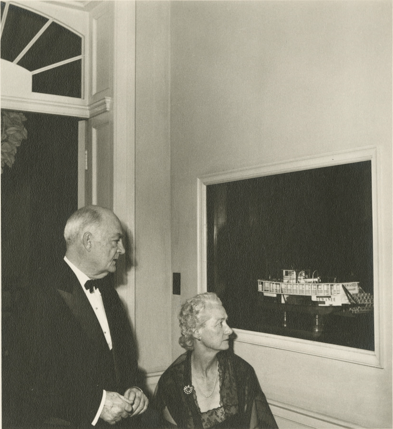 An elegantly dressed man and woman in formal attire admire a painting of a boat in a dimly lit room, with an arched window design in the background.