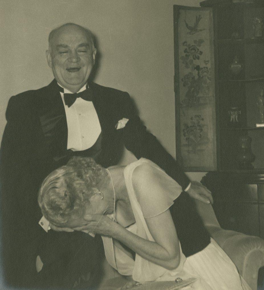 A man in a tuxedo stands next to a seated woman in a dress who is laughing and covering her face with her hands. They are in a room with ornate furnishings and a cabinet in the background.
