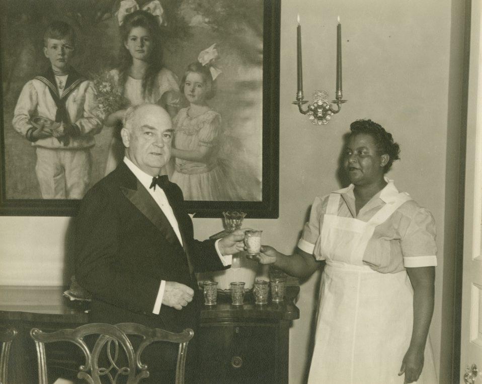 A formally dressed man receives a drink from a woman in a maids uniform in an elegant room. A large portrait of two children in vintage attire hangs on the wall behind them.