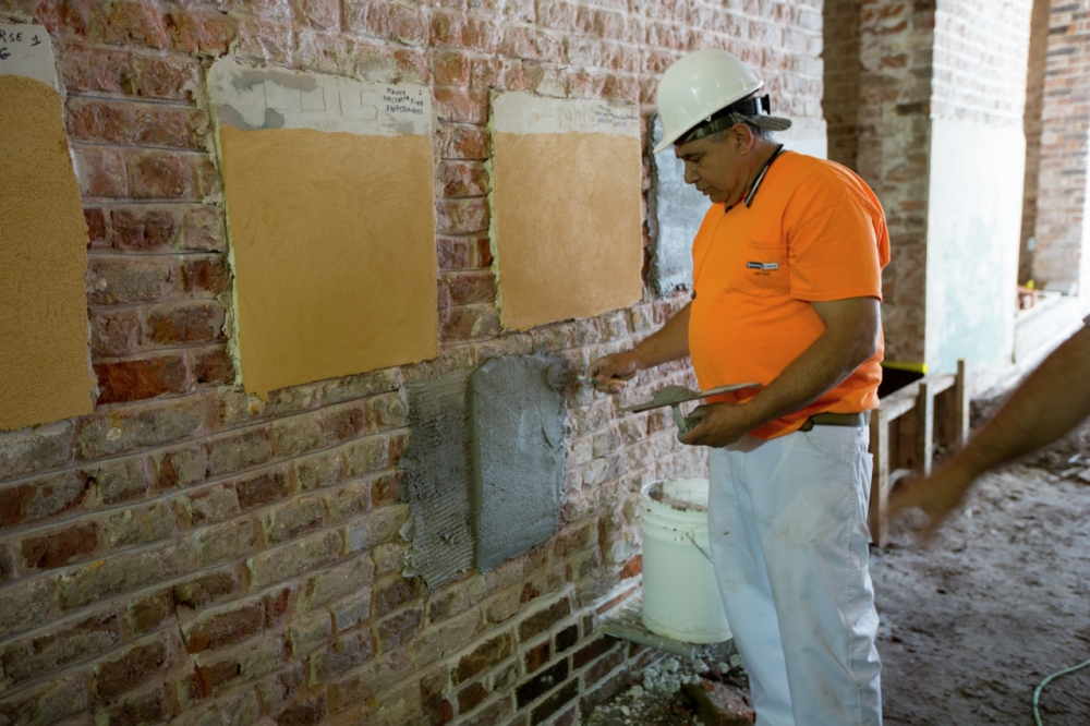 A construction worker in an orange shirt and hard hat applies cement to a brick wall using a trowel. Patches of different textures are visible on the wall. A white bucket and another workers arm are seen nearby in the unfinished room.