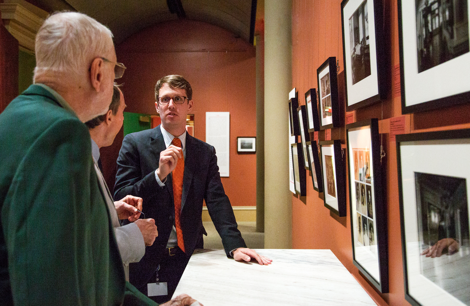 A man in a suit and glasses is speaking to three people in an art gallery. They are looking at a wall with framed photographs. The setting is formal, and the group appears engaged in discussion.