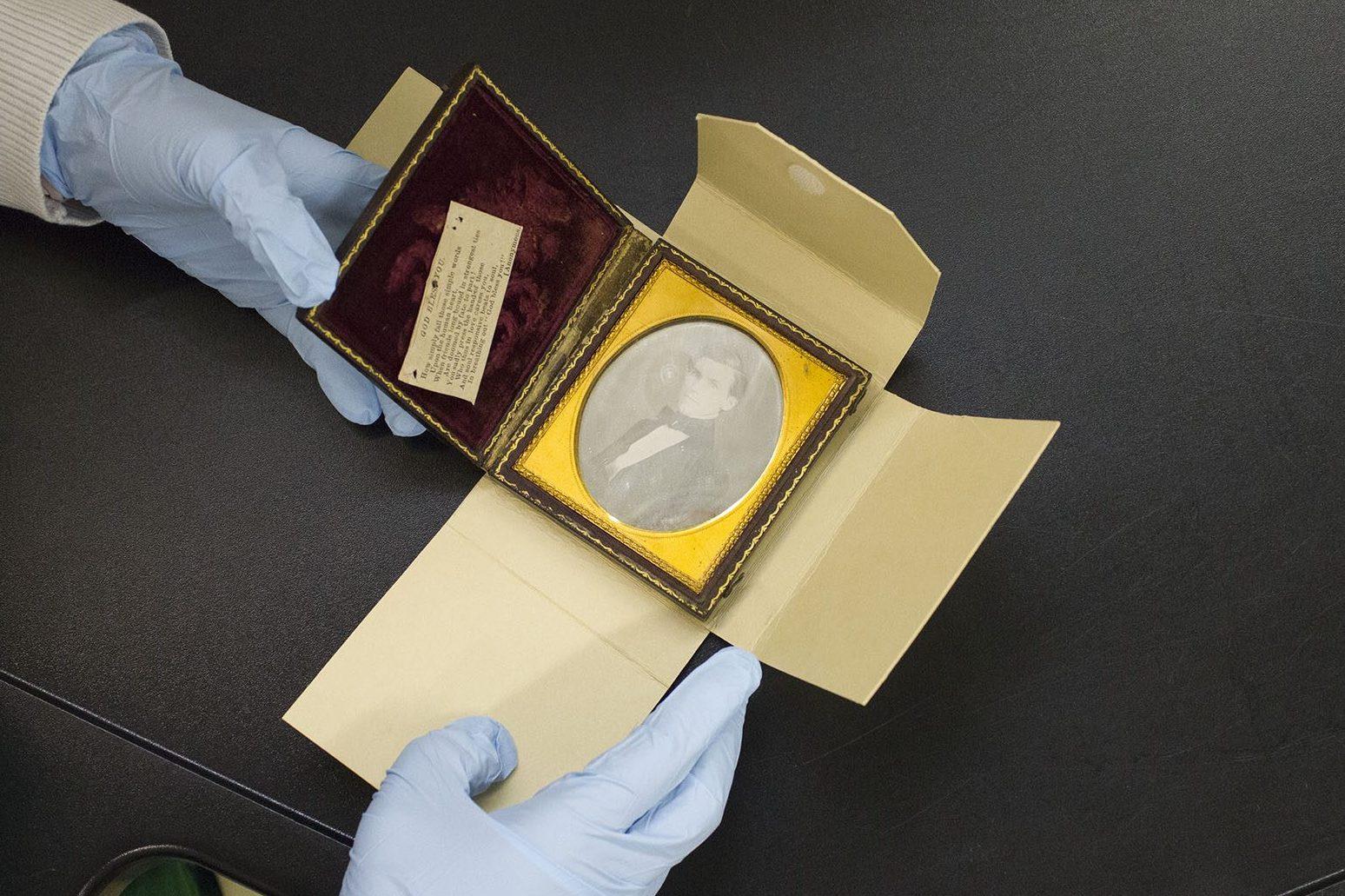 Gloved hands holding an opened antique photograph case with a vintage portrait and a label inside, resting on a black surface.
