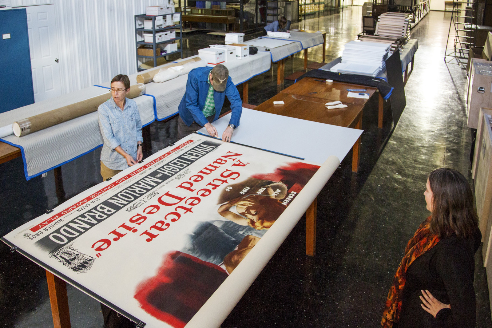 Three people handle a large vintage poster on a table in a spacious room filled with various paper materials and equipment. The poster features bold text and a classic design. The scene suggests a conservation or restoration process.